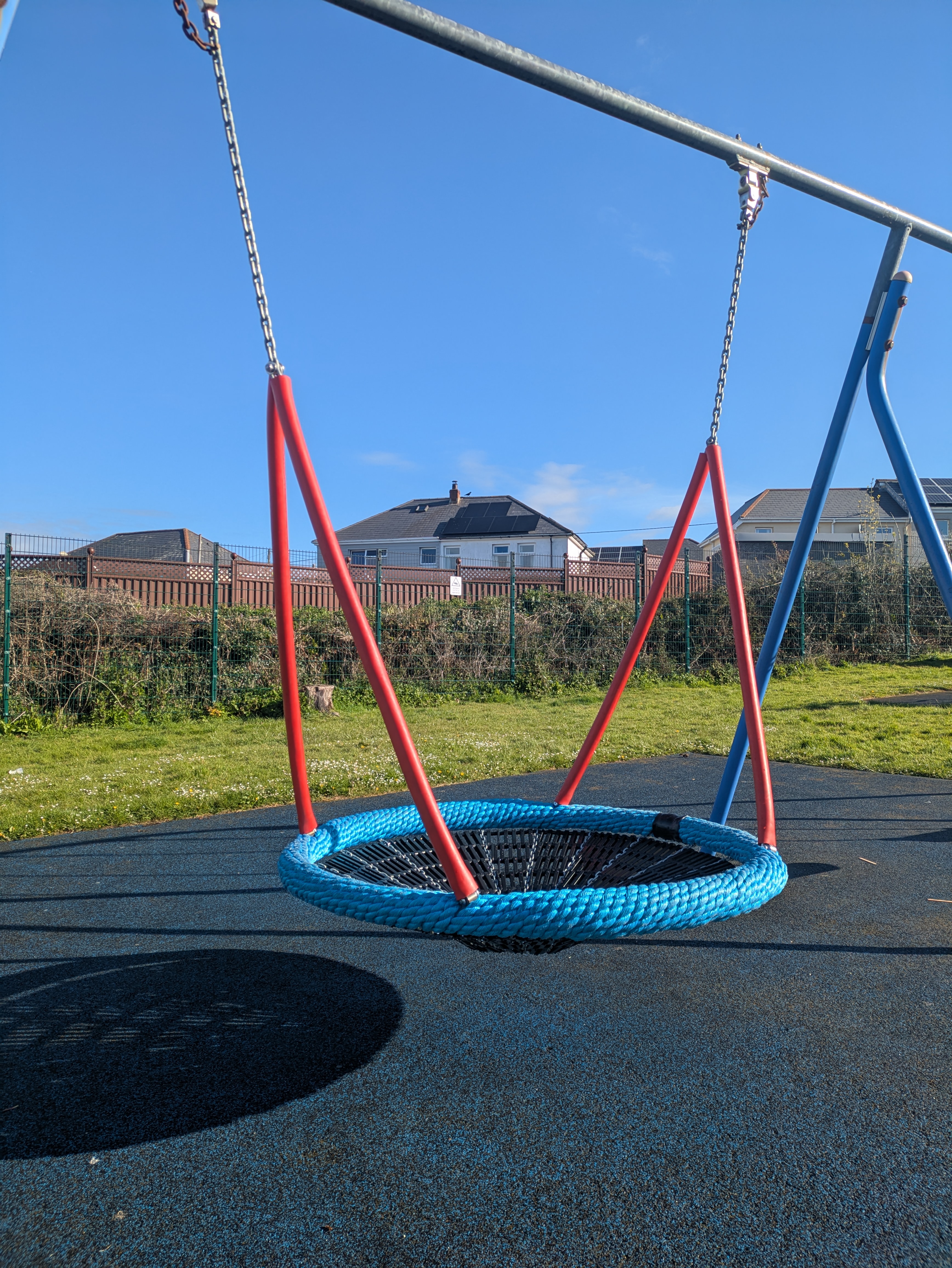 Sun shining on a new red and blue basket swing, with green grass, roof tops and clear blue sky behind.